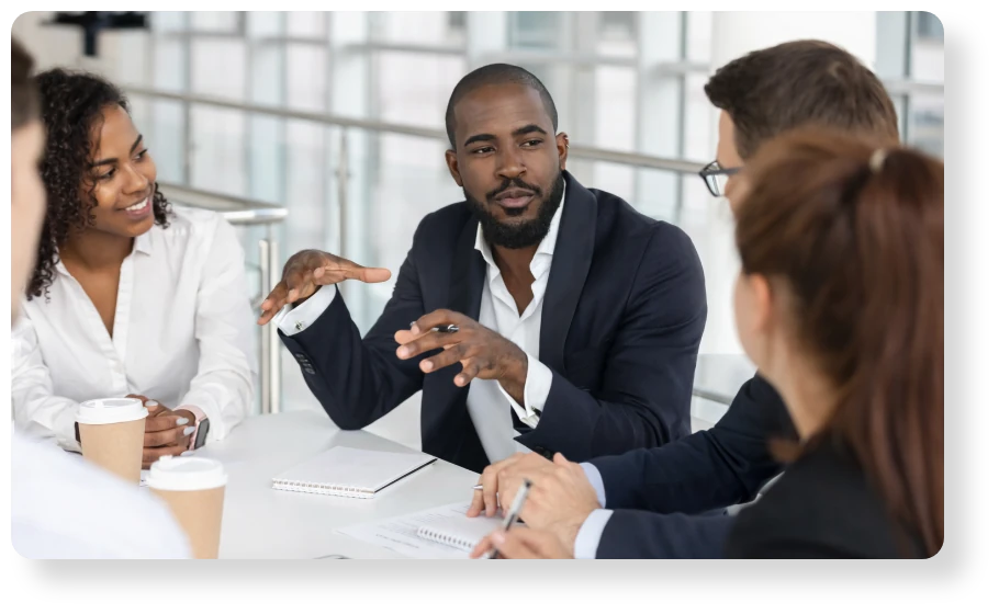 Business professionals gathered around a table, with one man leading a discussion and gesturing as colleagues listen.