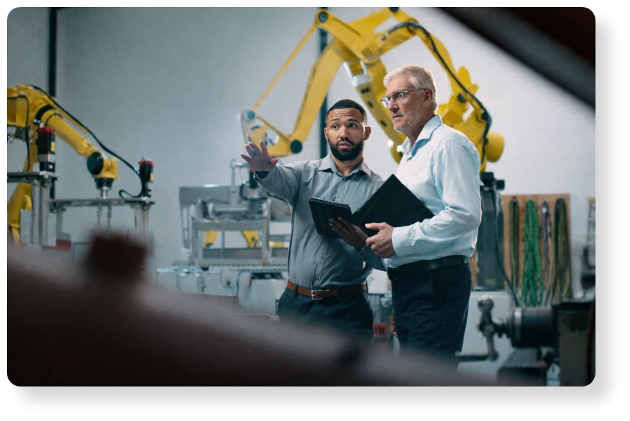 Two engineers discuss operations in a modern manufacturing facility, with industrial robotic arms in the background.