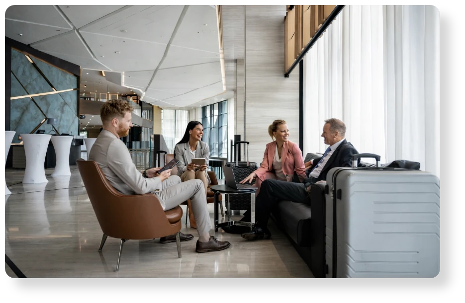 Four business professionals having a conversation in a modern airport lounge, with luggage and large windows in the background.