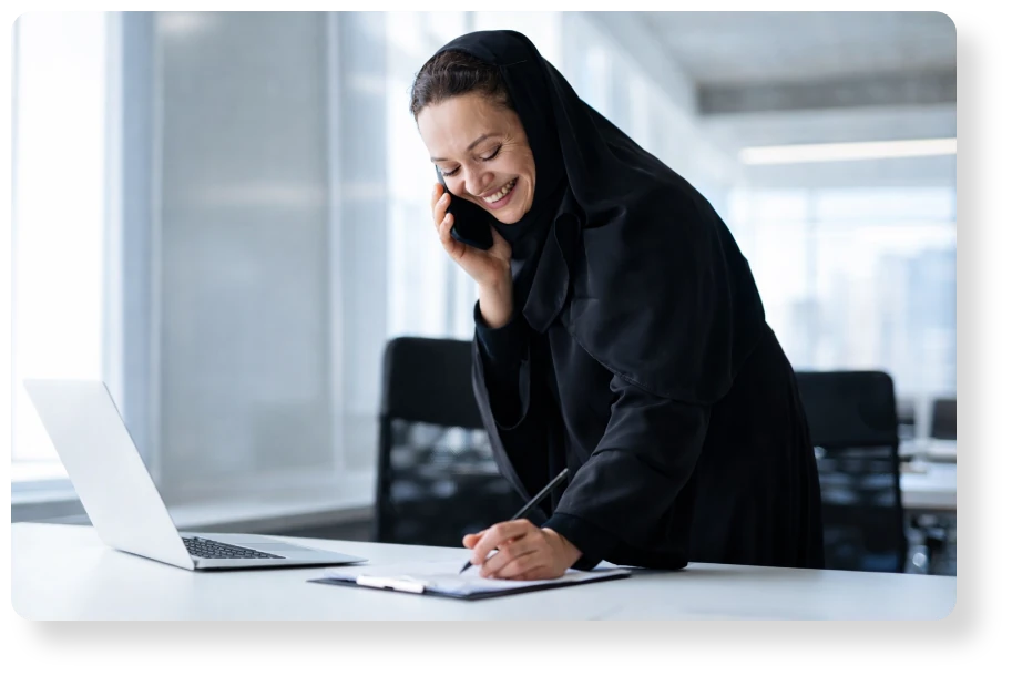Smiling professional wearing a black hijab, speaking on the phone while taking notes beside a laptop in a modern office.