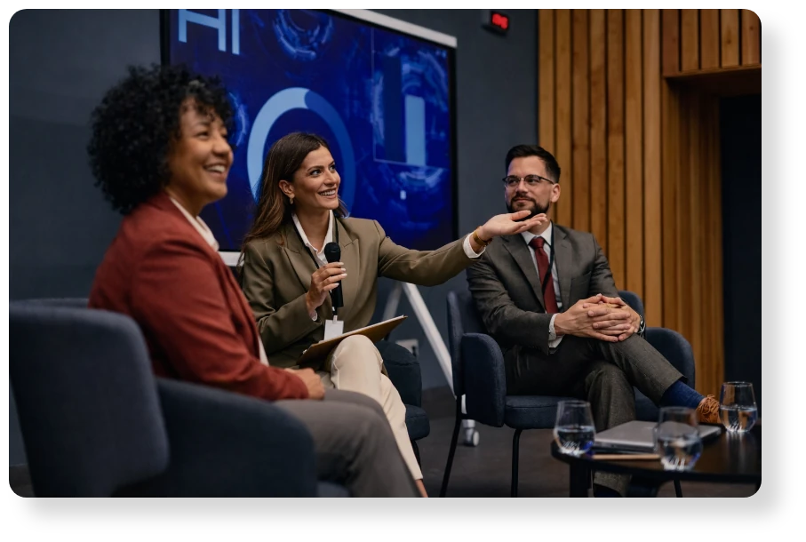 Three business professionals seated in a modern discussion setting, with one woman speaking into a microphone and gesturing during a panel conversation.