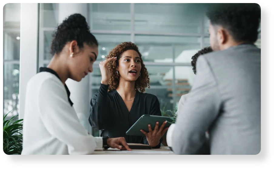 A professional leads a discussion with colleagues, holding a tablet and sharing ideas during a collaborative office meeting.