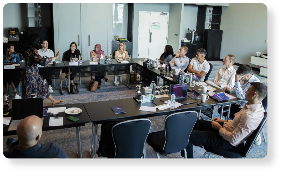 A diverse group of professionals gathered around tables in a modern meeting room, listening and discussing during a collaborative workshop.