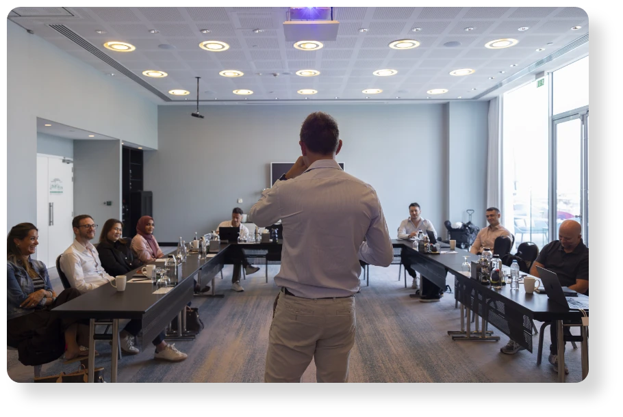 A team leader addresses colleagues seated around tables during a meeting in a modern office.